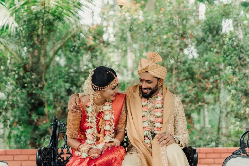 Shreya & Sanketh laughing together during post-wedding shoot while sitting on metal two-seater chair with outdoor lighting